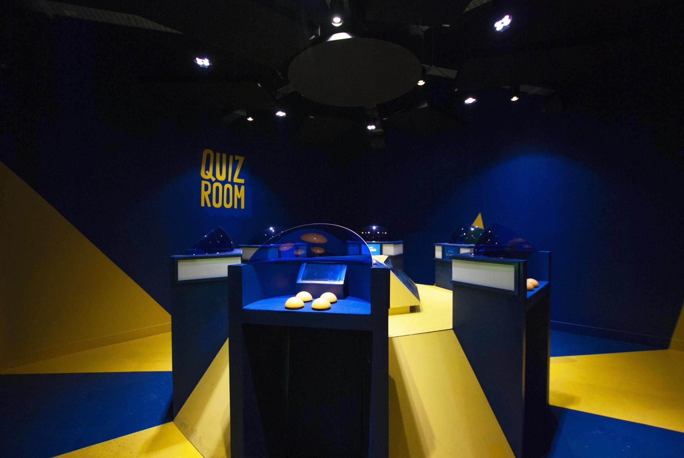 Une salle de Quiz Room aux couleurs bleu et jaune, avec ses pupitres de jeu